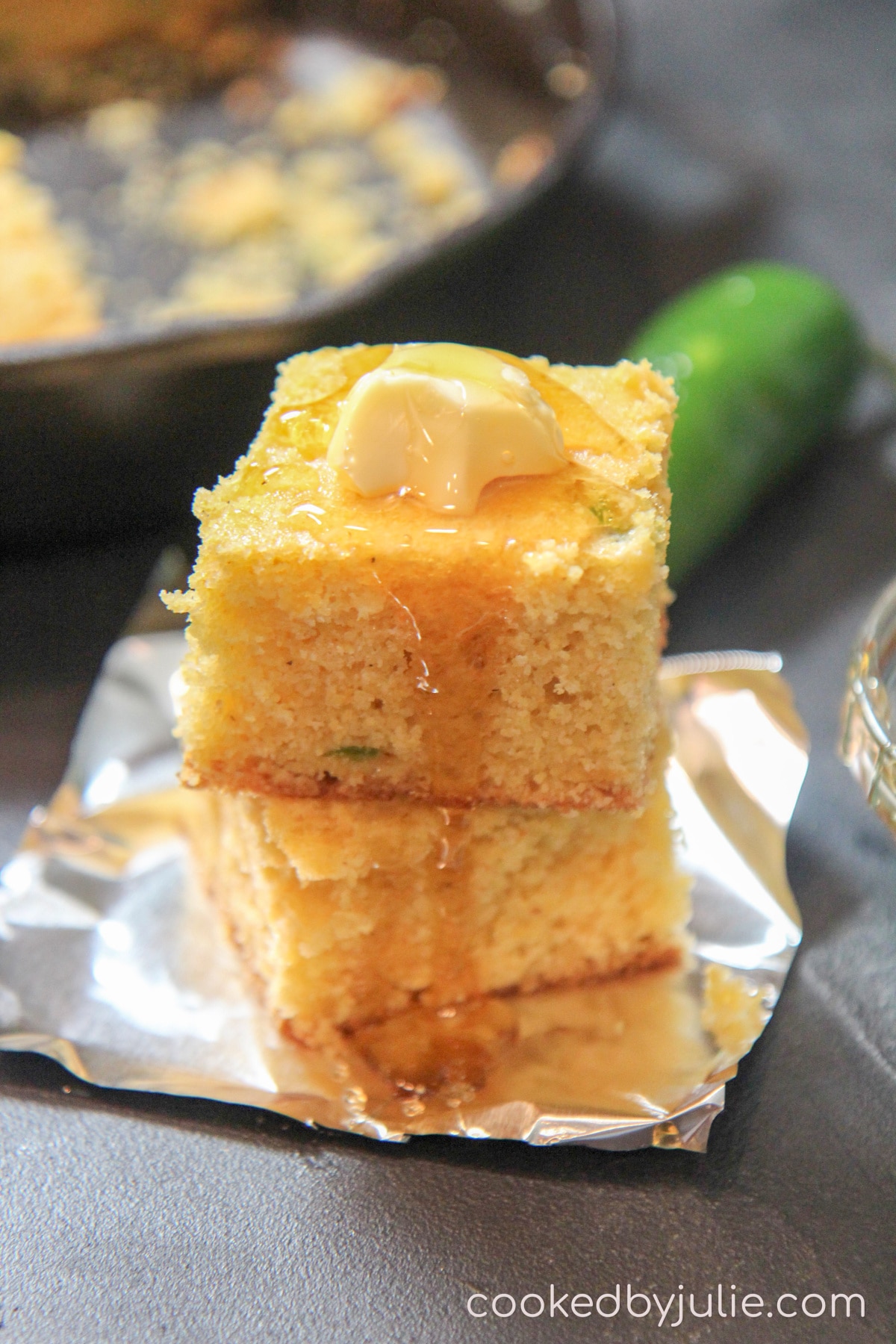 two squared honey cheddar jalapeno cornbread slices on top of aluminum foil with a jalapeno pepper in the back ground and an iron skillet. 