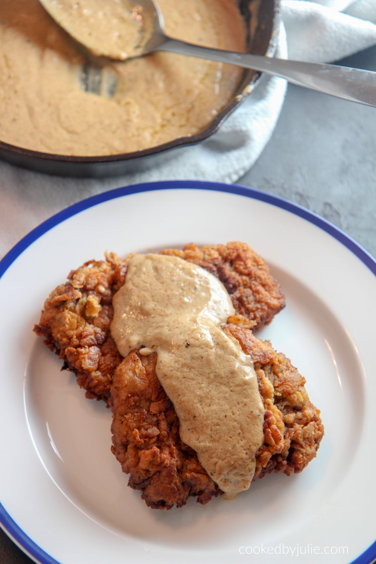 two chicken fried steak cutlets with gravy on top and an iron skillet with gravy and a spoon in the background. 
