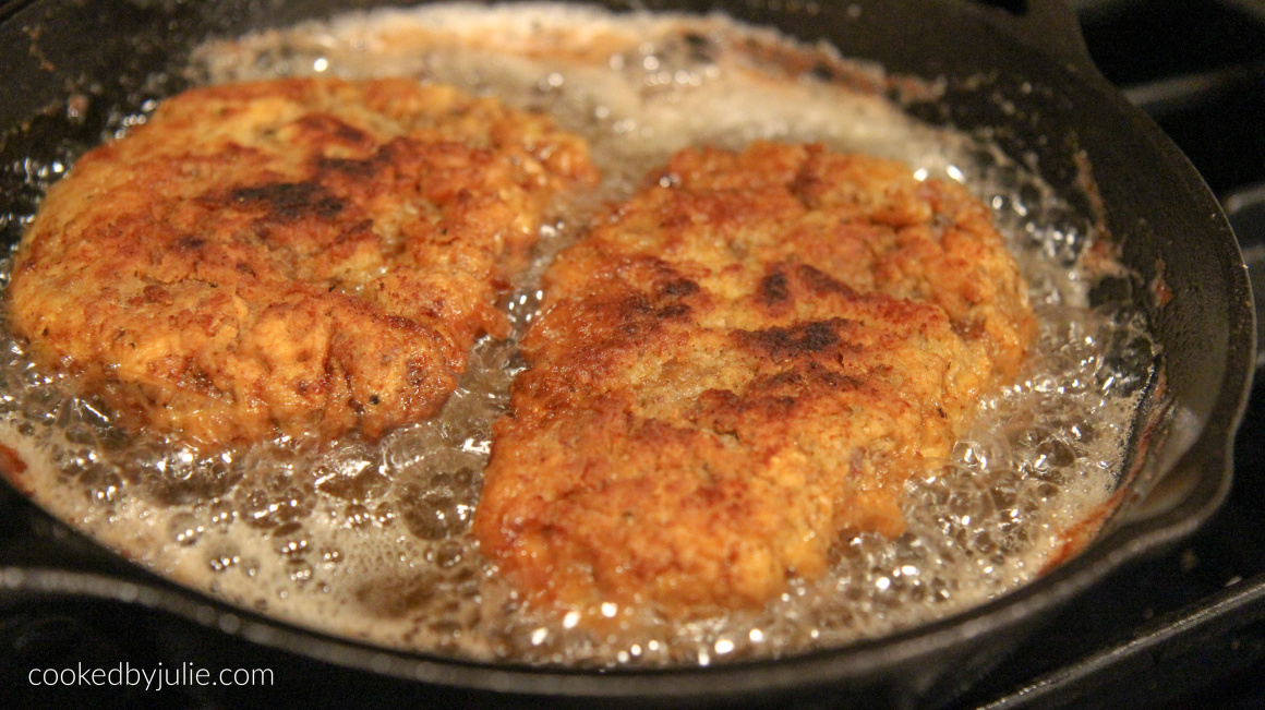 two chicken fried steak cutlets frying in an iron skillet with oil. 