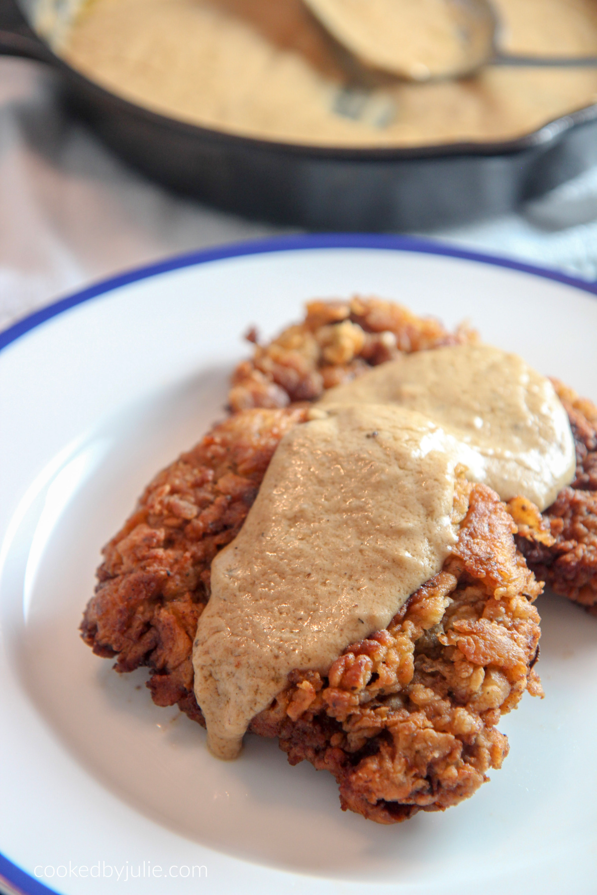 two pieces of chicken fried steak on a white and blue plate with gravy on top. An iron skillet with gravy and a spoon in the background. 