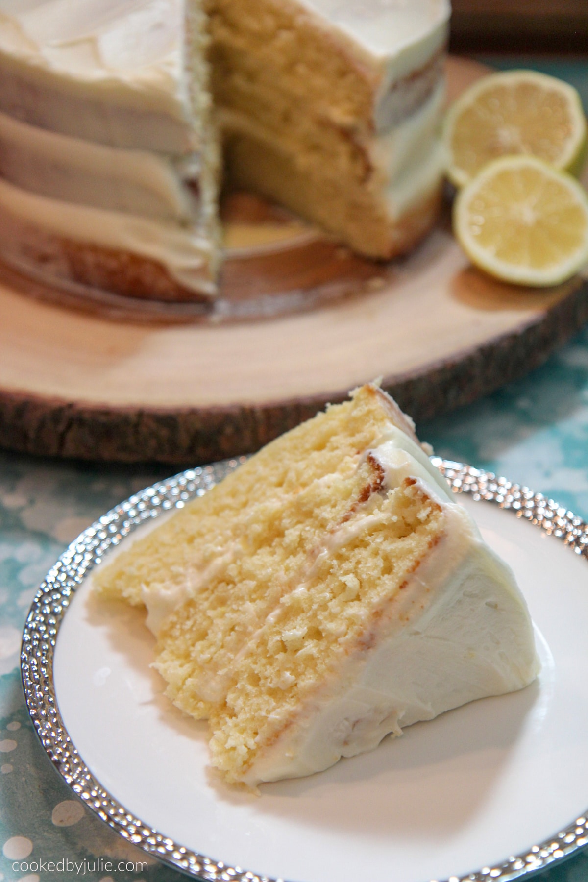 a lemon cake slice on a white and silver plate and a lemon cake in the background on a wooden cake stand with 2 lemon slices. 
