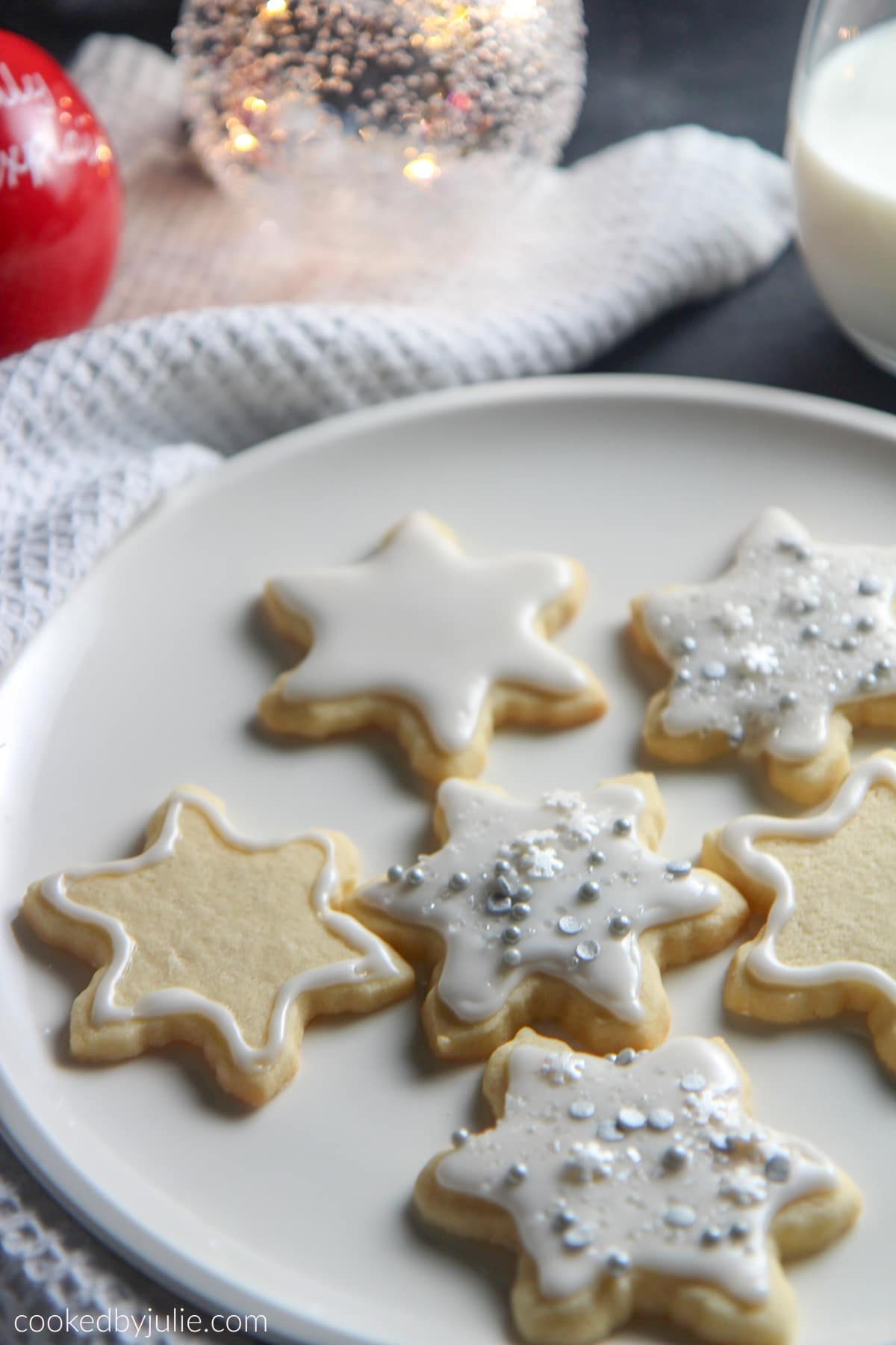 six cookies on a white plate with a white towel on the side, a glass of milk, and two Christmas tree balls on the side. 