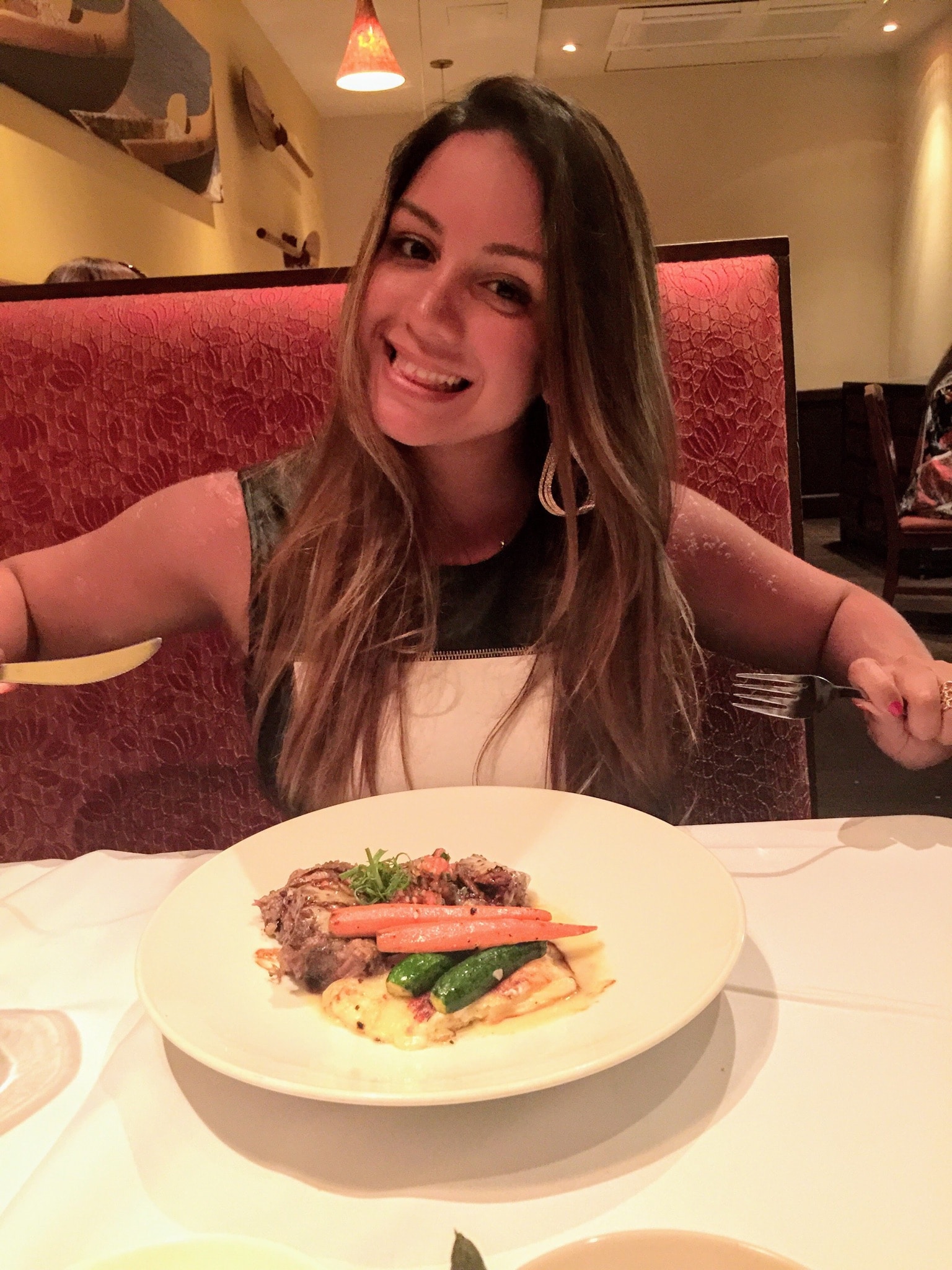 A female with long hair at a restaurant booth with a fork and knife and plate of food.