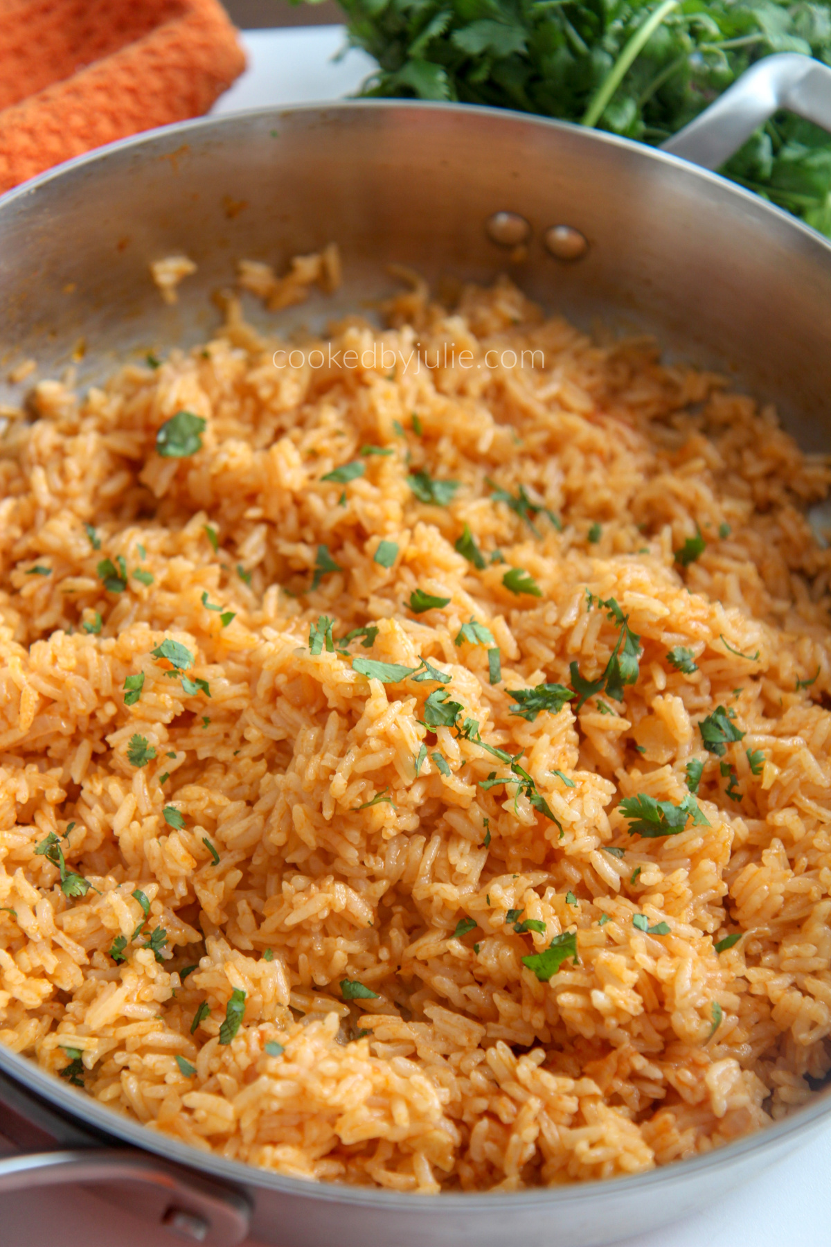 A skillet filled with Spanish rice with an orange kitchen towel and fresh cilantro in the background