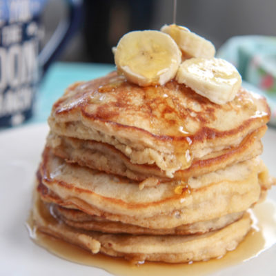 six banana pancakes on a white plate with banana slices on top, a drizzle of maple syrup, and a blue mug in the background.
