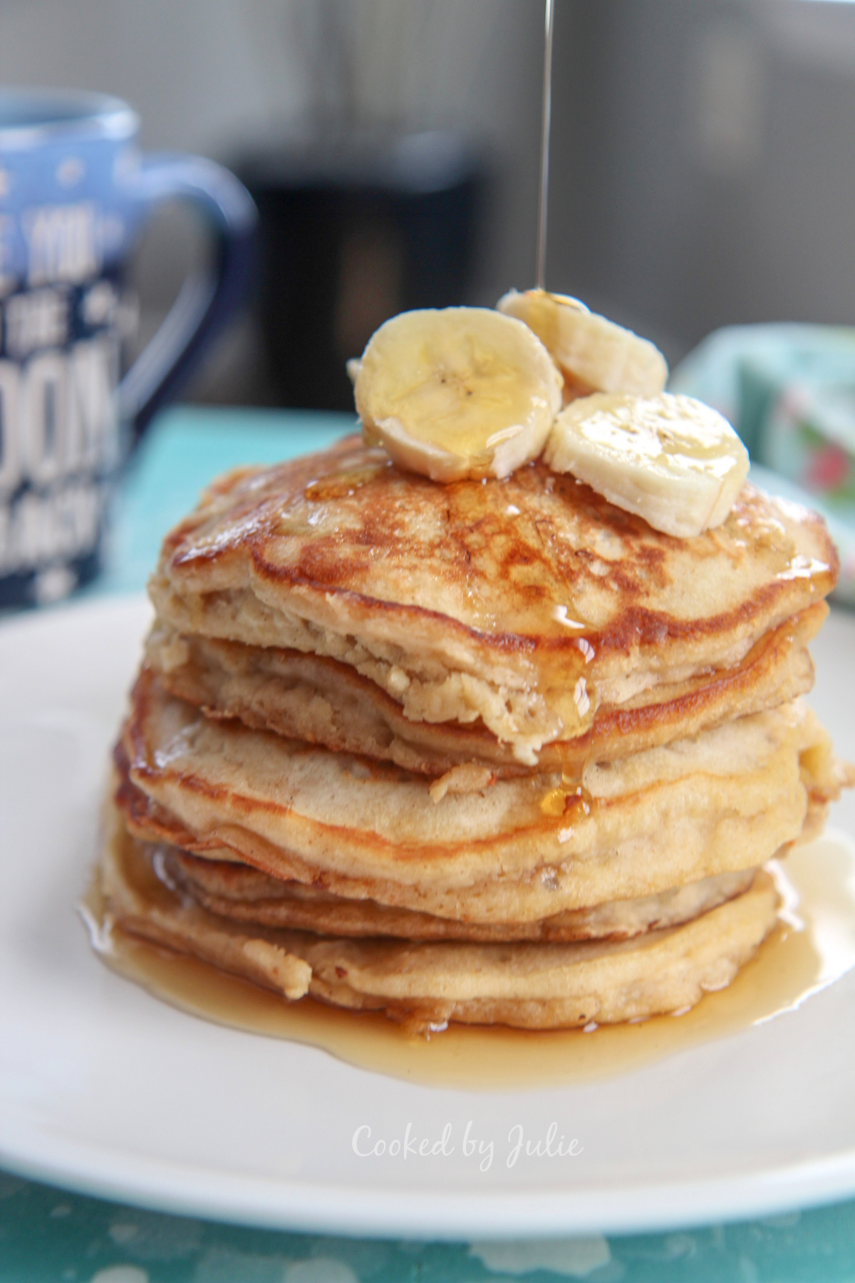 six banana pancakes on a white plate with banana slices on top, a drizzle of maple syrup, and a blue mug in the background.