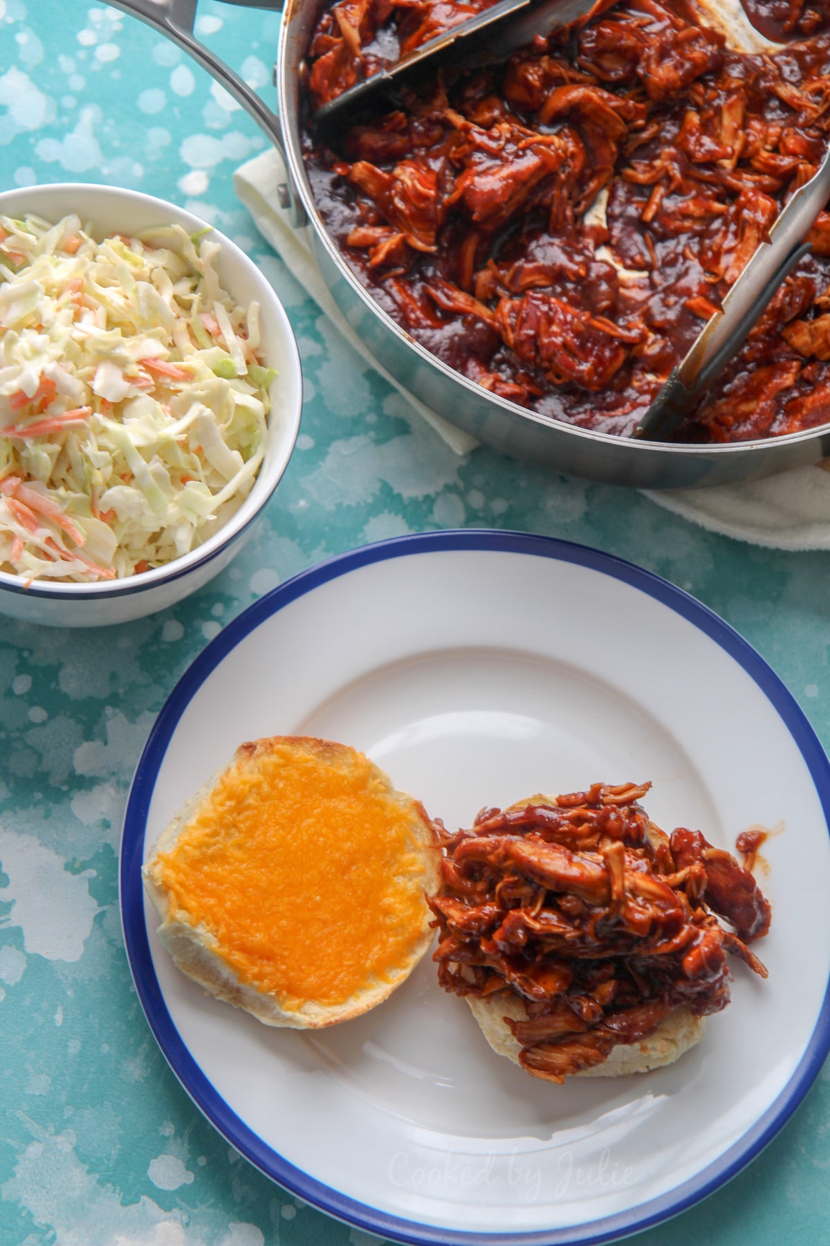 pulled chicken sandwich with cheddar cheese on a white and blue plate. Coleslaw in a bowl and pulled chicken in a skillet