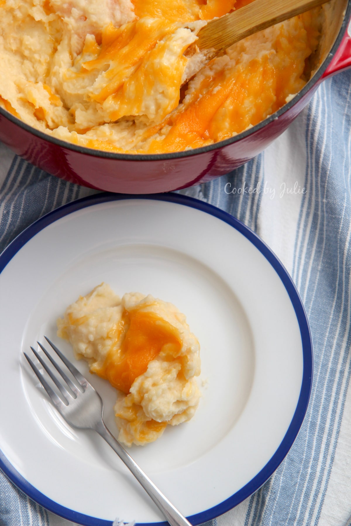 white and blue plate of cheddar mashed potatoes with a fork and red pot filled with mashed potatoes