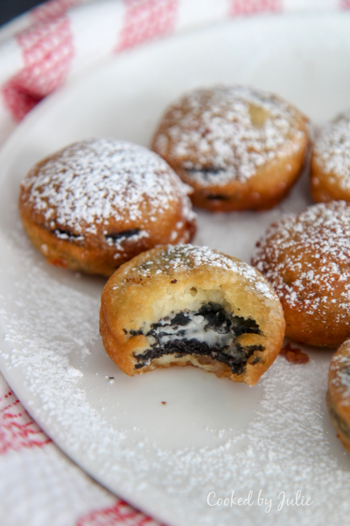 deep fried Oreos with powdered sugar on a white plate with one half eaten cookie and a red and white towel on the side. 
