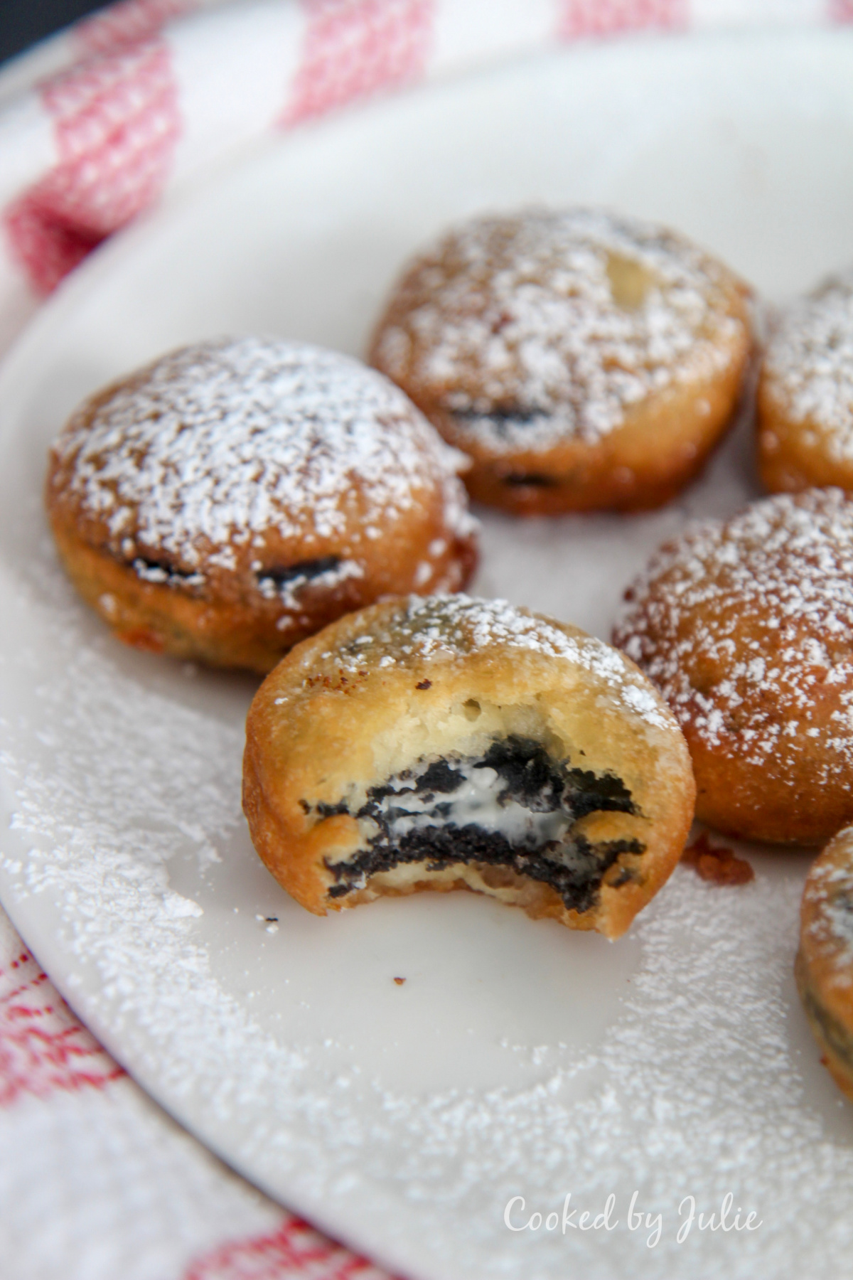 deep fried Oreos with powdered sugar on a white plate with one half eaten cookie and a red and white towel on the side.