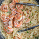 shrimp over fettuccine alfredo in a black skilletw ith tongs and fresh parsley in a black skillet.