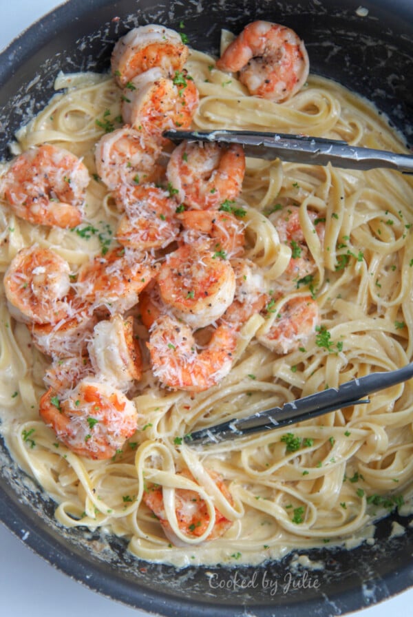 shrimp over fettuccine alfredo in a black skilletw ith tongs and fresh parsley in a black skillet.