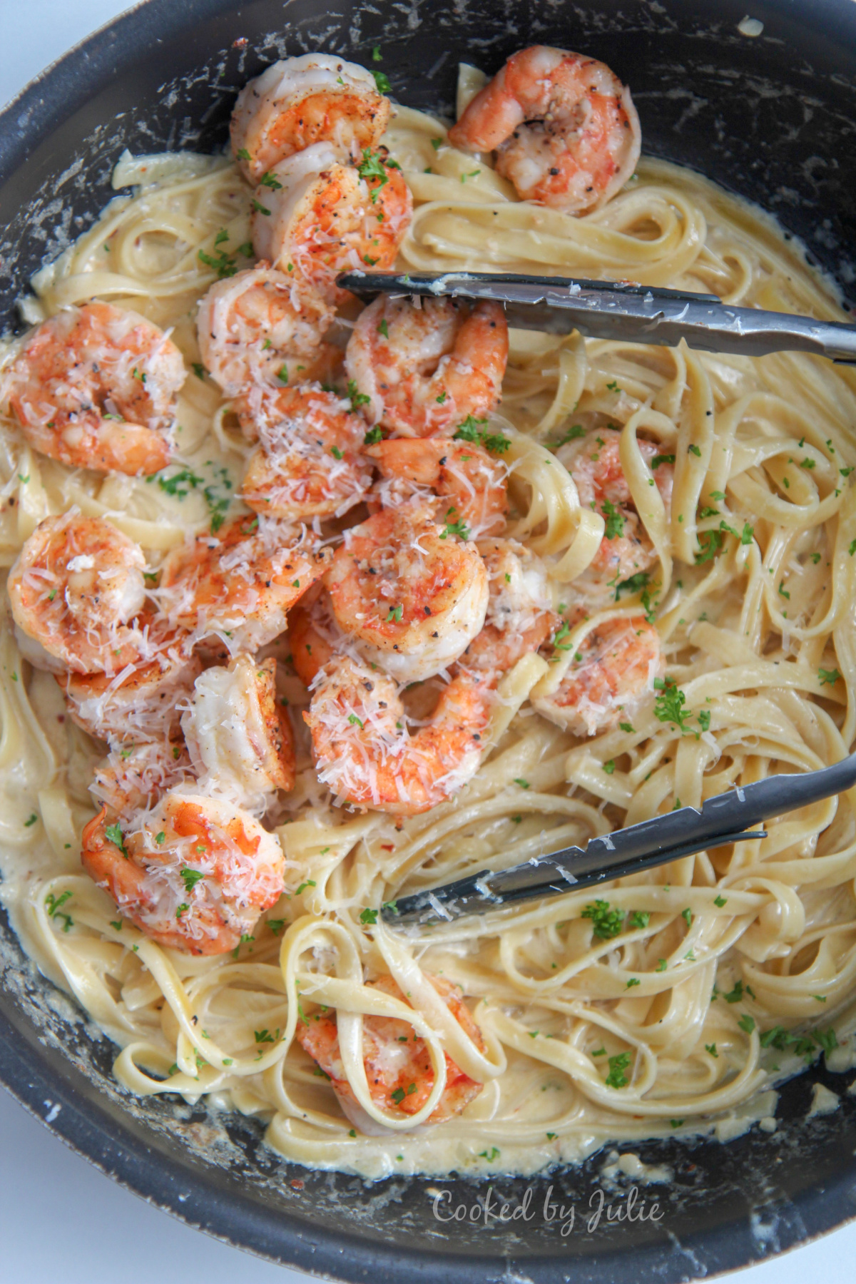 shrimp over fettuccine alfredo in a black skillet with tongs and fresh parsley 