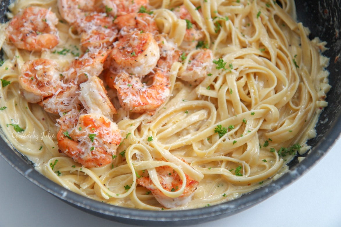 shrimp fettuccine alfredo with fresh parmesan and parsley in a black skillet on top of a white table