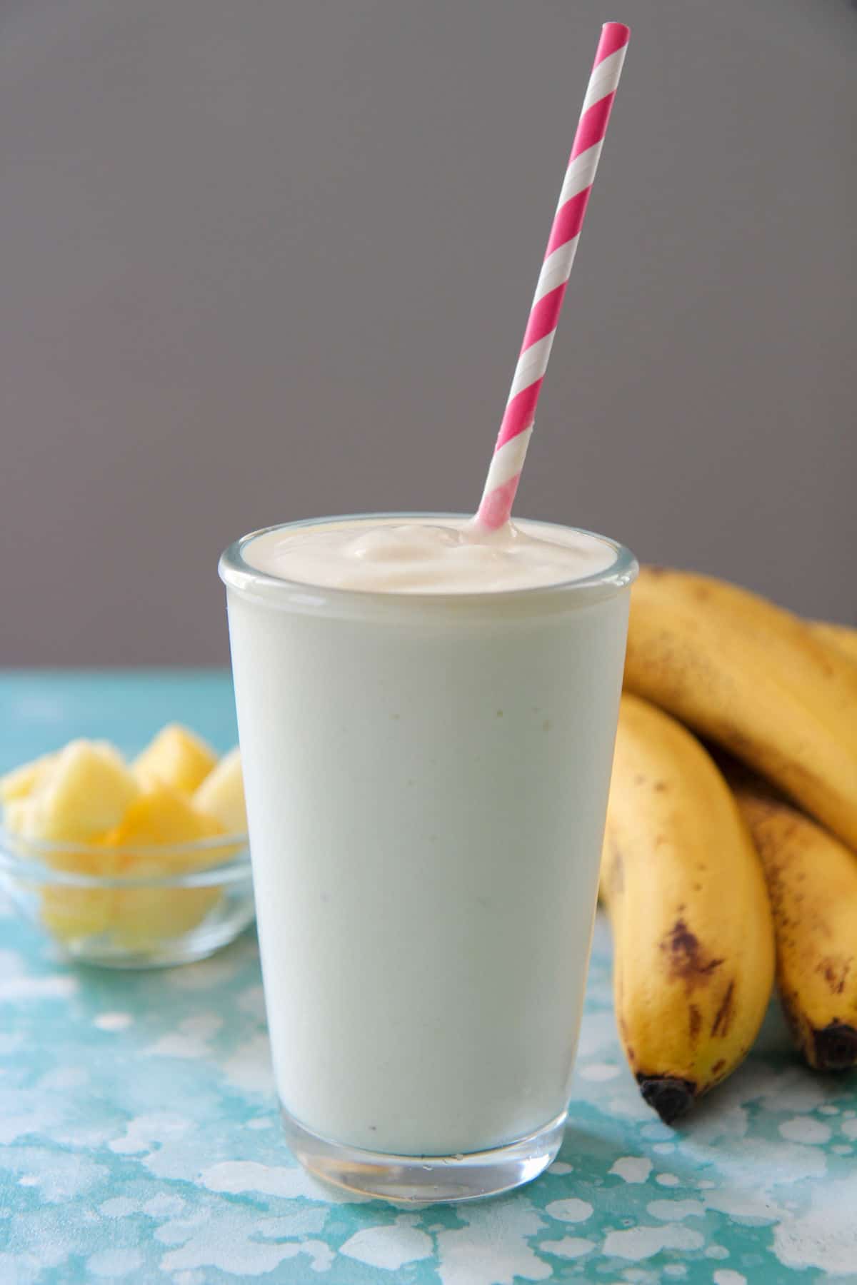 pineapple smoothie in a tall glass with a pink and white straw and bananas and pineapples in the background. 