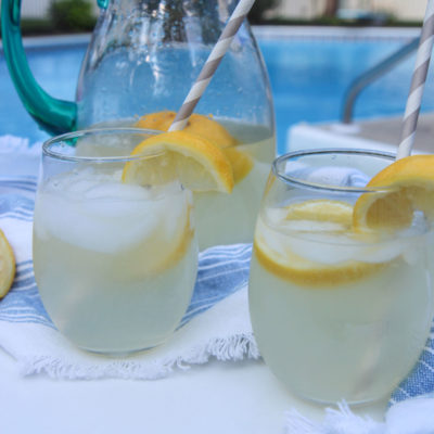 two glasses of lemonade and a glass pitcher in the background