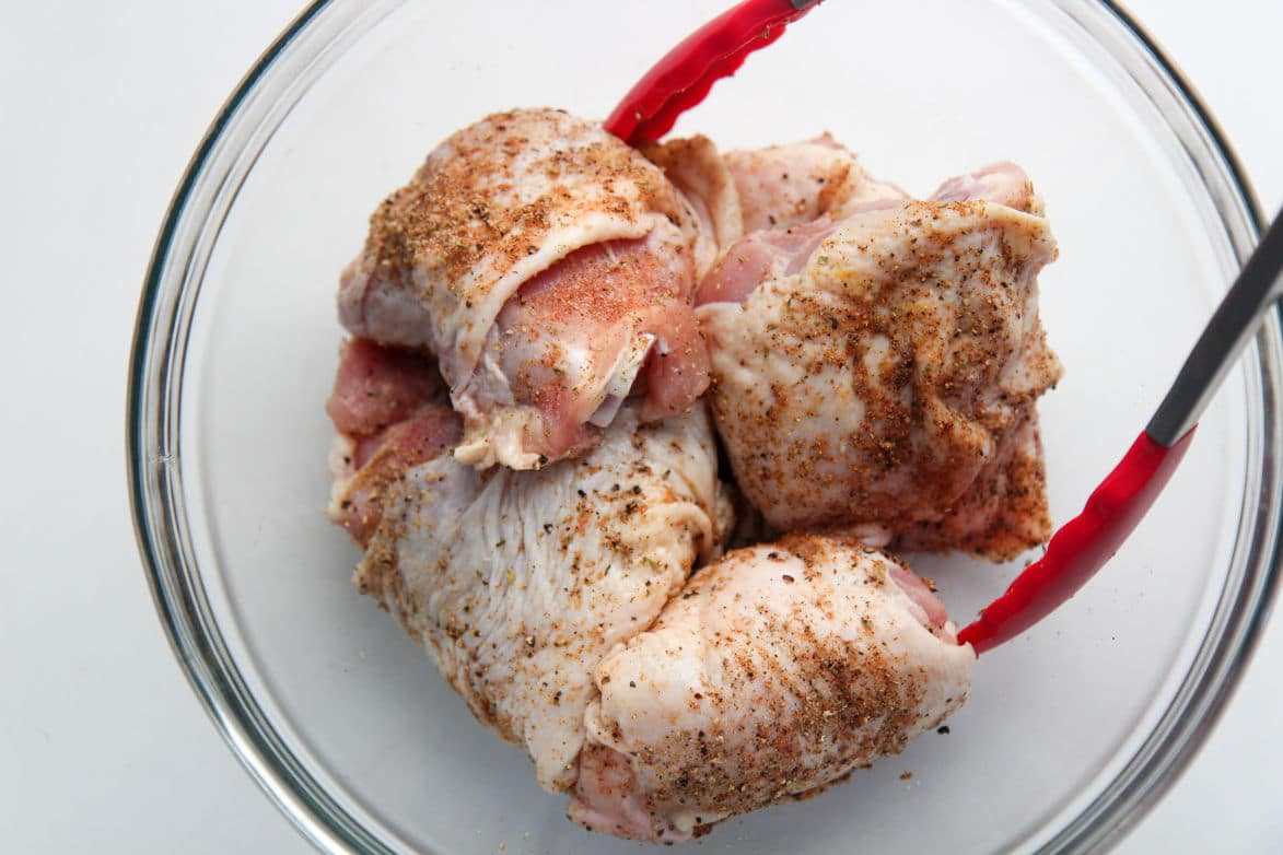 seasoned chicken thighs in a glass bowl with red tongs 