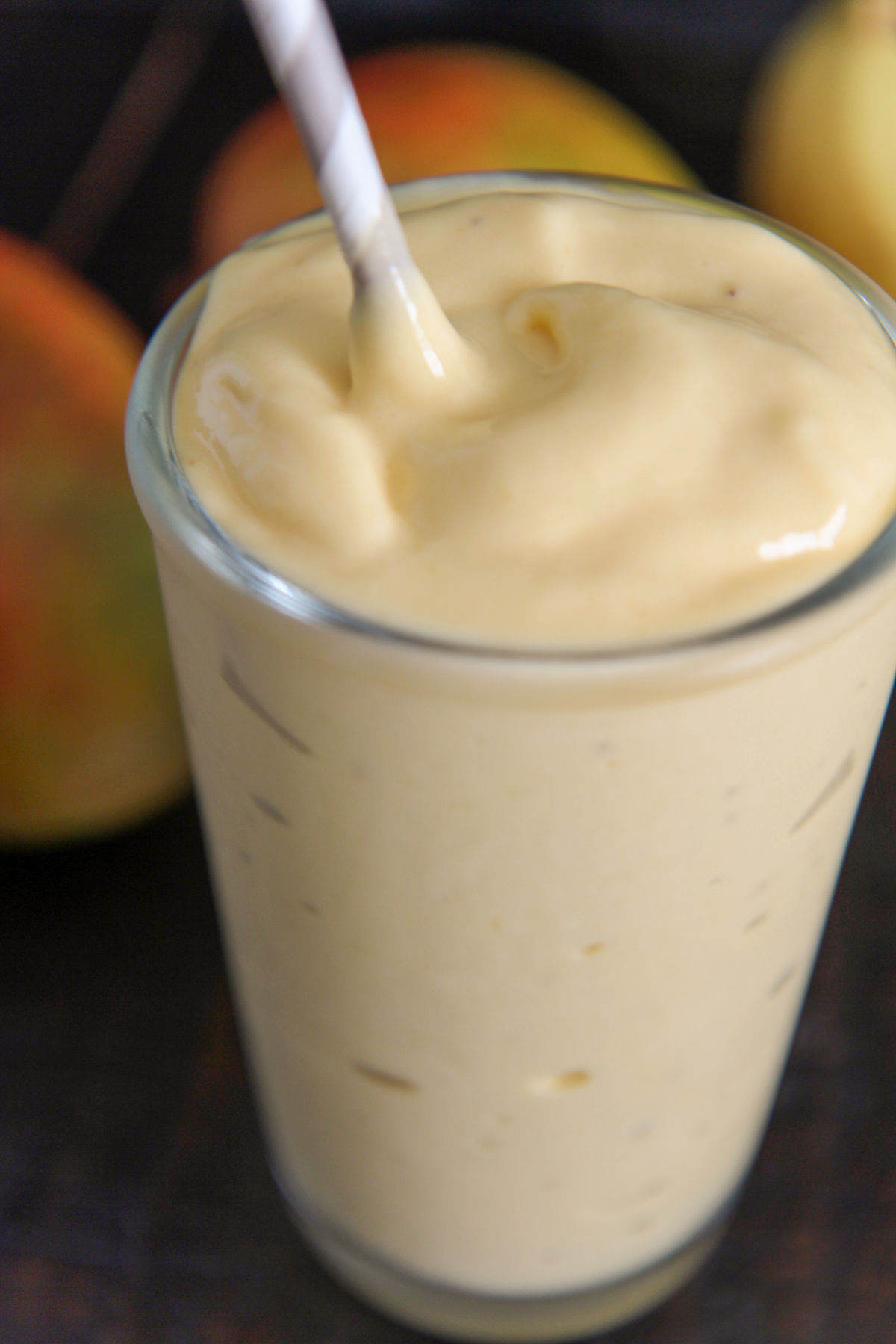 mango smoothie in a glass with a white straw and mangoes in the background 