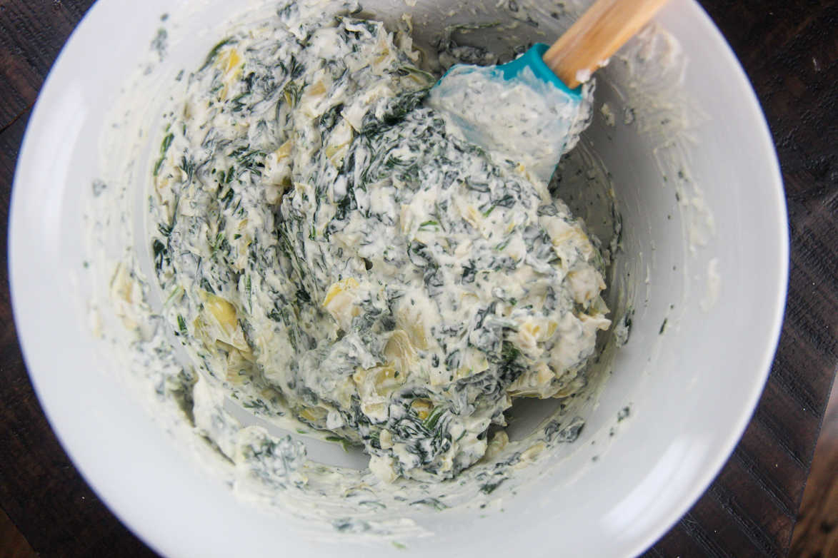 spinach artichoke dip mixture in a white bowl with a blue spatula 