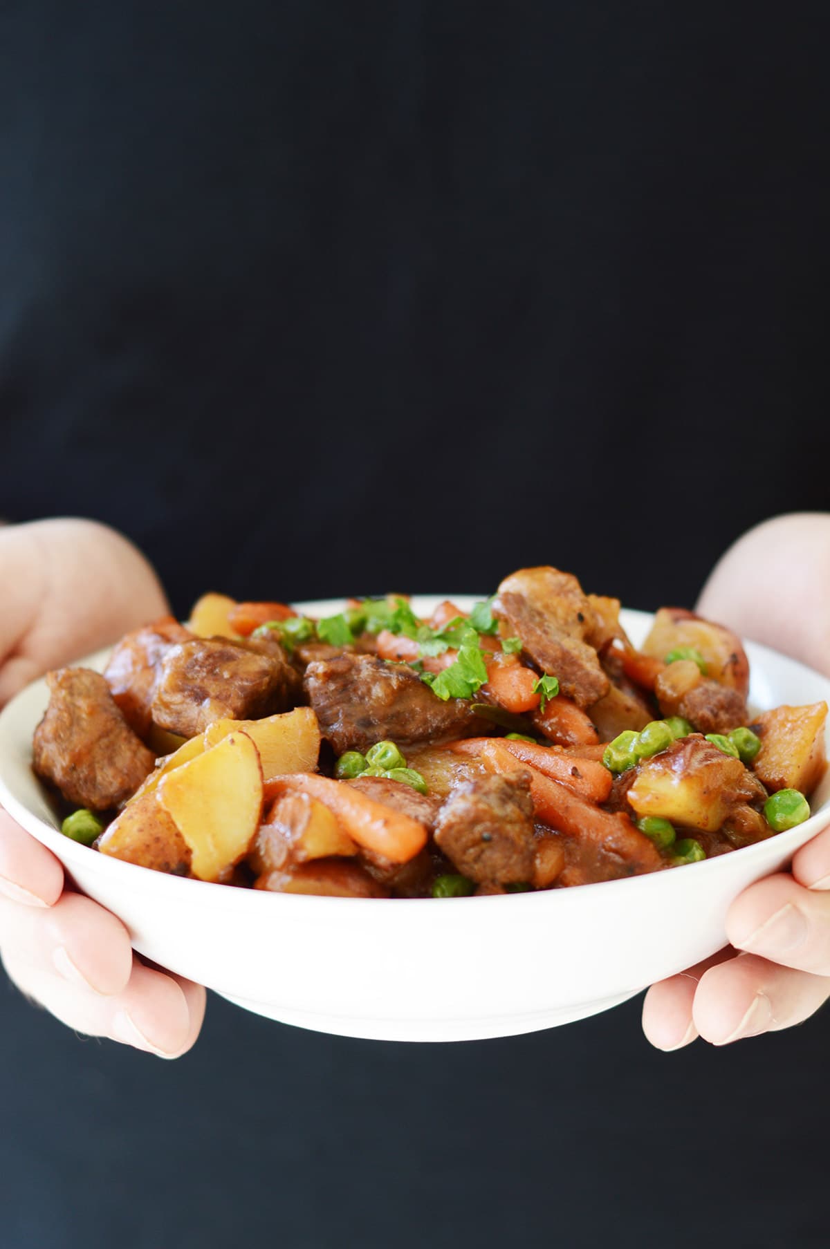 two hands holding a white bowl of meat, potatoes, and carrots.