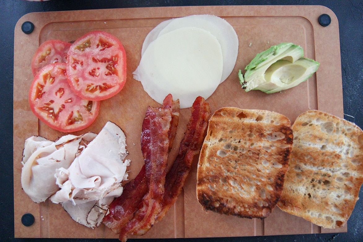 tomatoes, bacon, turkey, cheese, avocado, and bread on a wooden board