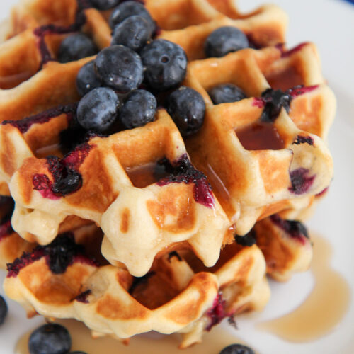 two buttermilk blueberry waffles on a white plate with maple syrup and fresh blueberries.