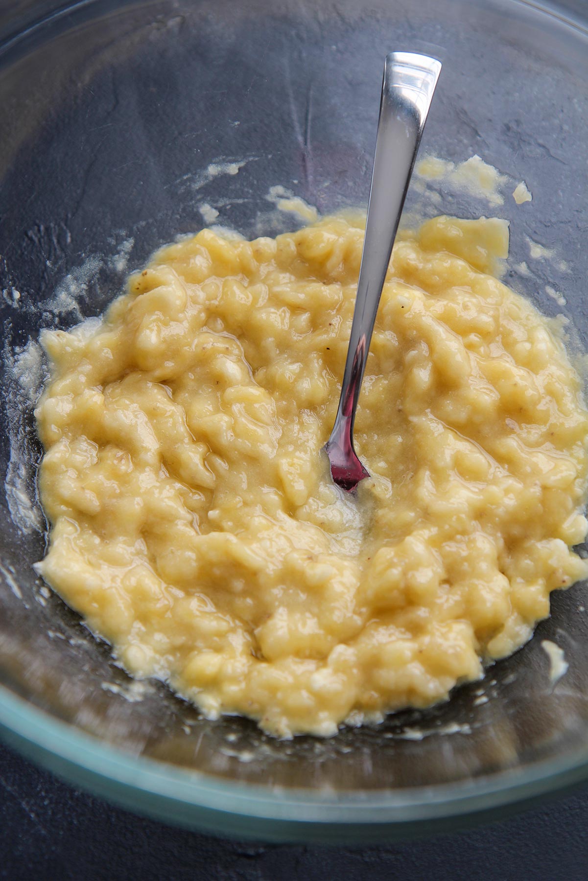 four bananas mashed in a glass bowl with a fork on a dark surface.