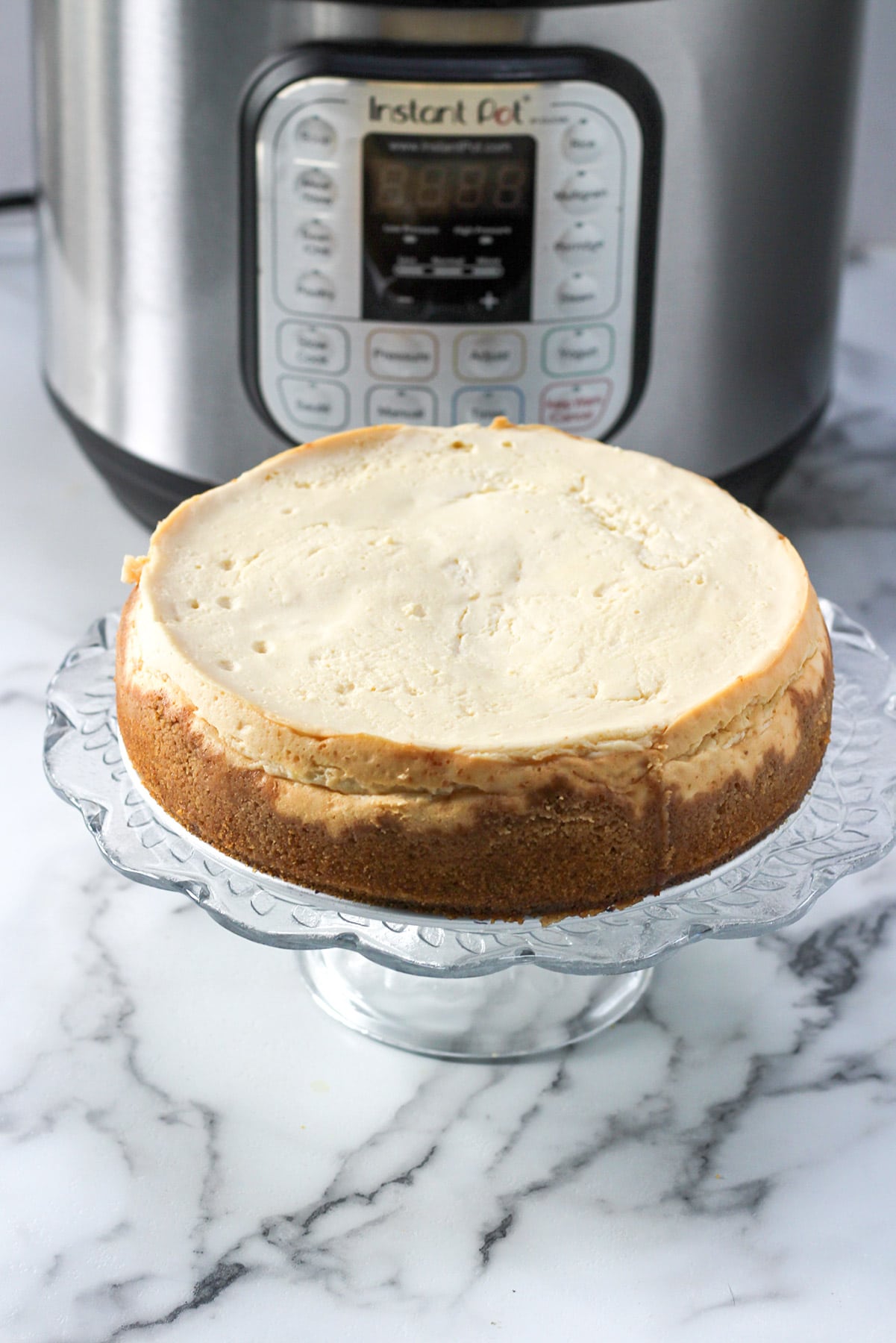 plain cheesecake on a glass stand and a pressure cooker in the background. 