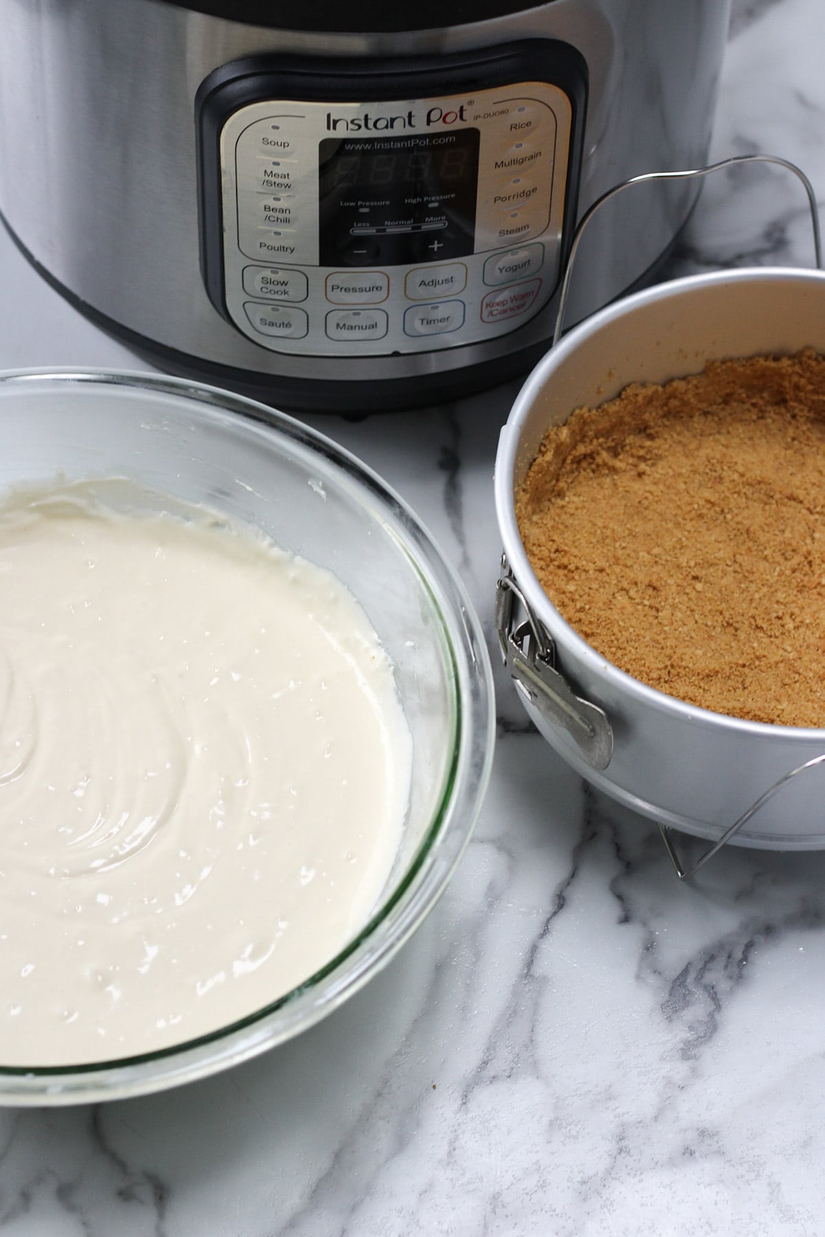 cheesecake batter in a glass bowl, a spring form pan with a graham cracker crust and an instant pot in the background. 