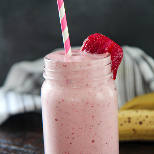 strawberry banana milkshake in a mason jar with fresh strawberries, a straw, and bananas in the background.