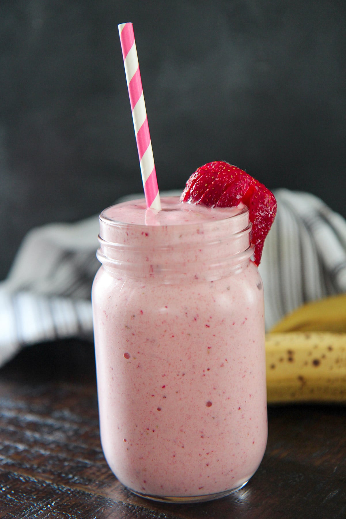 strawberry banana milkshake in a mason jar with fresh strawberries, a straw, and bananas in the background. 