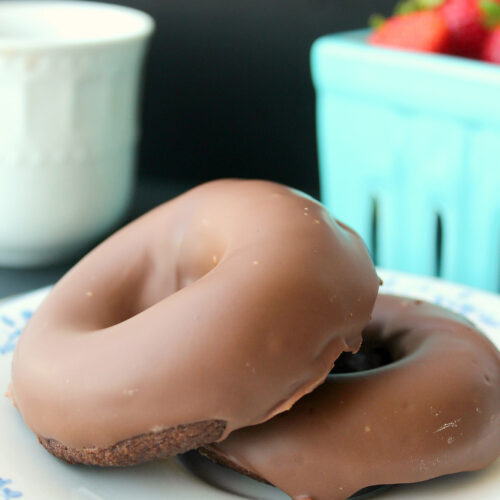 two keto baked chocolate donuts on a white plate with a basket of strawberries in the background.