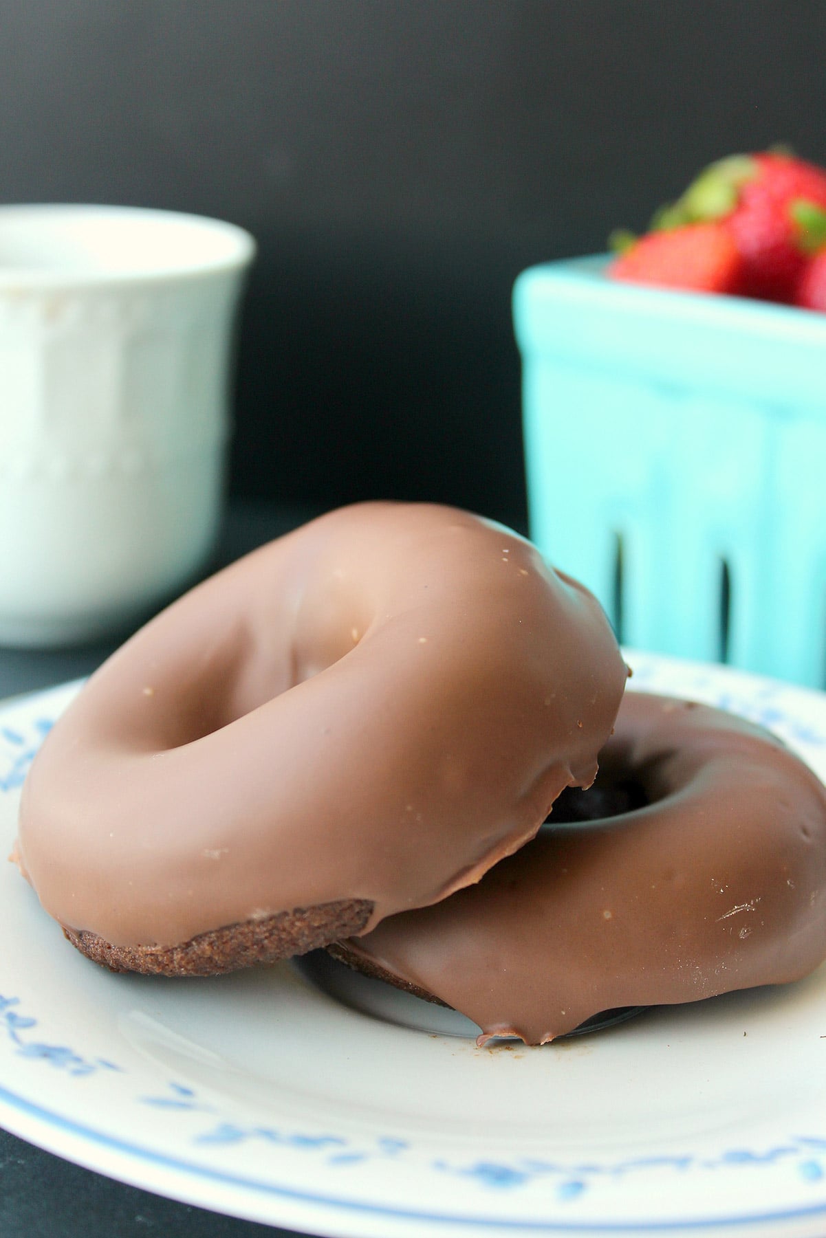 two keto baked chocolate donuts on a white plate with a basket of strawberries in the background.