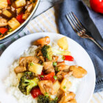 pineapple chicken stir-fry over white rice on a white plate with a fork on the side and skillet in the background.