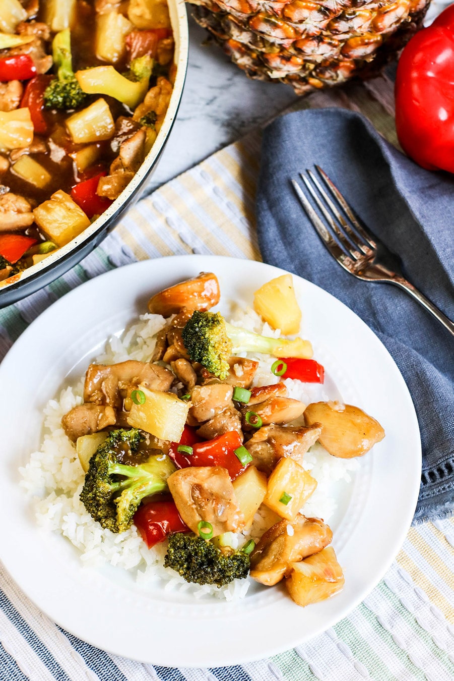 pineapple chicken stir-fry over white rice on a white plate with a fork on the side and skillet in the background. 