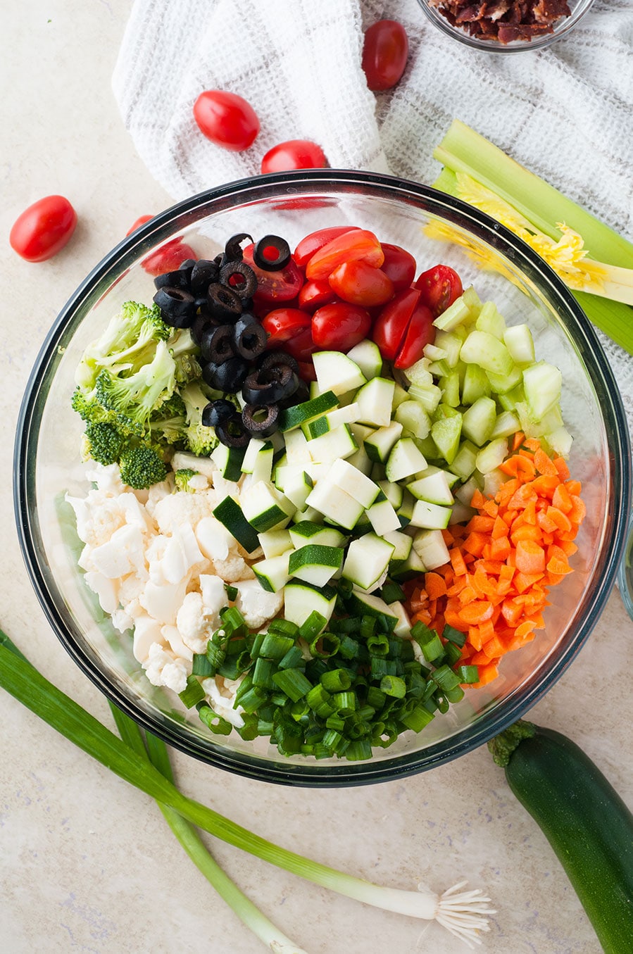 a large bowl filled with carrots, zucchini, broccoli, black olives, tomatoes, and cauliflower. 