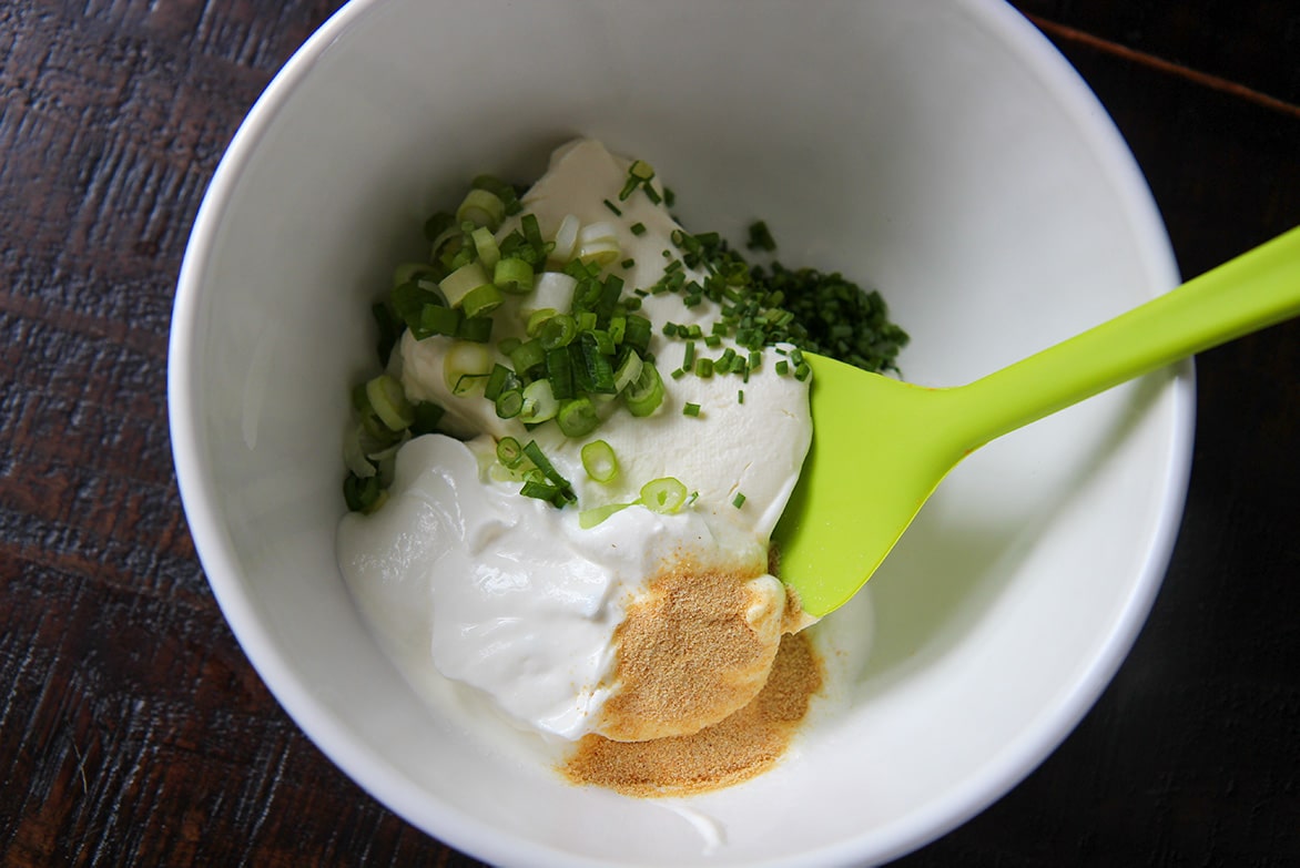 cream cheese, sour cream, granulated garlic, chives, scallions, and a green spatula in a white bowl. 