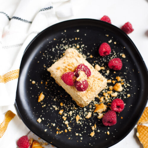 a squared slice of no-bake dalgona cheesecake on a black plate with raspberries and crushed cookies on top. A white and yellow towel on the side.