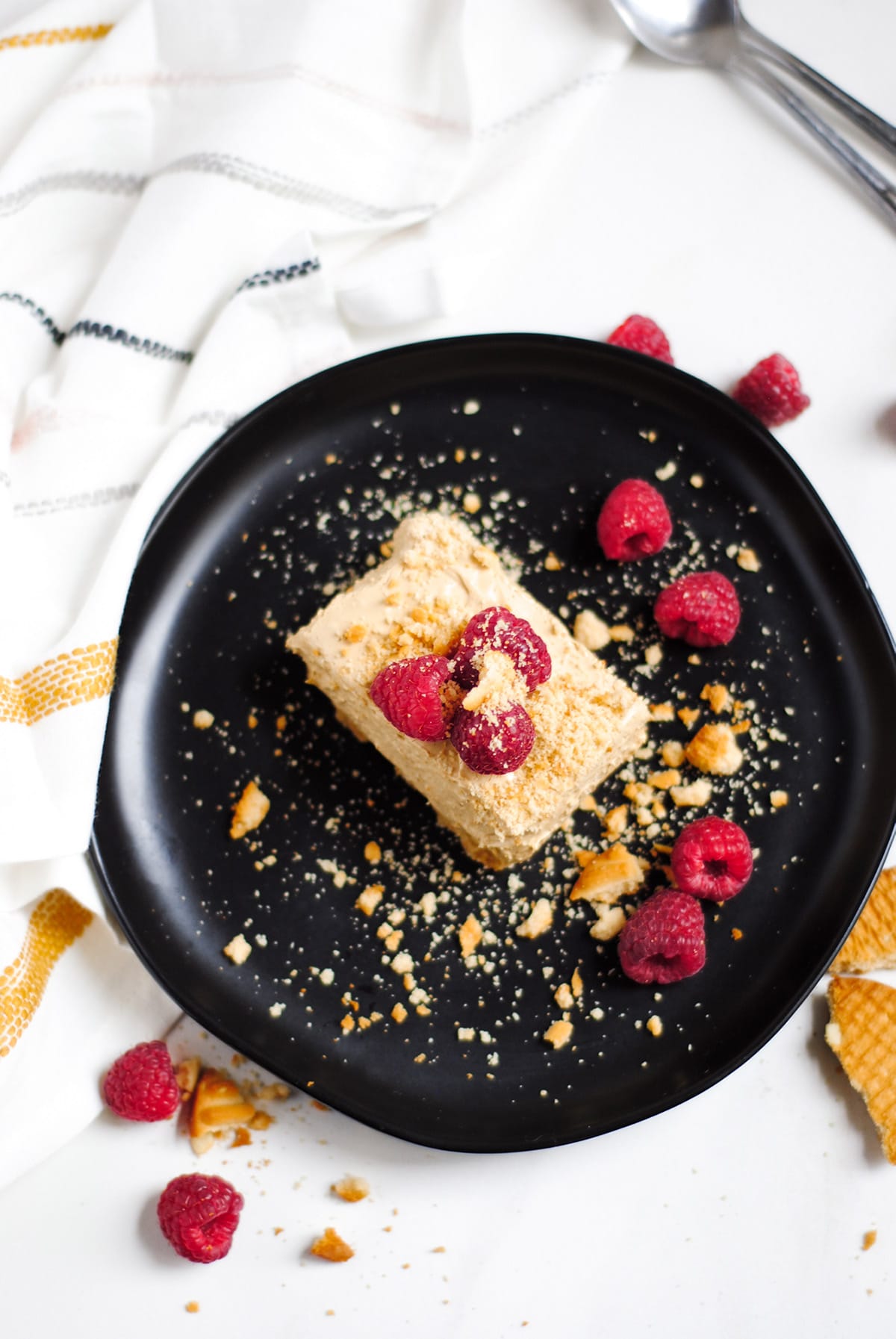 a squared slice of no-bake dalgona cheesecake on a black plate with raspberries and crushed cookies on top. A white and yellow towel on the side. 