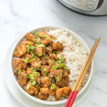 sesame chicken with a side of white rice in a white bowl with chop sticks on top and an instant pot in the background.