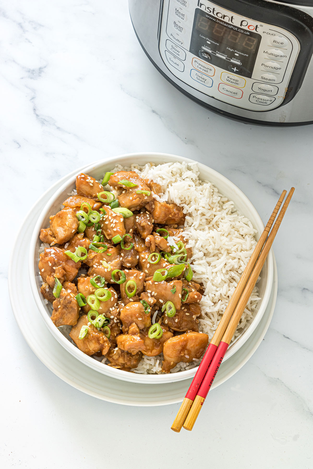 sesame chicken with a side of white rice in a white bowl with chop sticks on top and an instant pot in the background. 