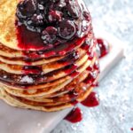 a stack of thin pancakes on a light surface with blueberry sauce on top of the pancakes.