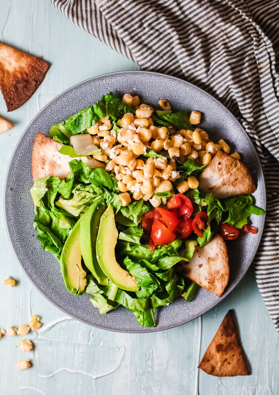 lettuce, corn, avocado, and tortilla chips in a blue bowl with a black and white striped towel on the side. 