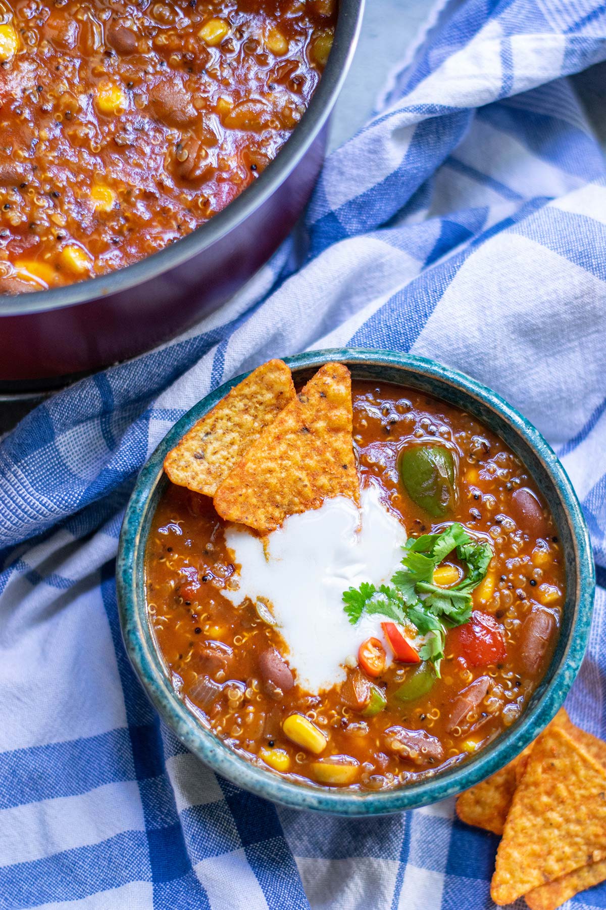 a small blue bowl filled with quinoa chili and dorito chips on the side and in the bowl. A pot with chili in the background and a blue towel underneath. 
