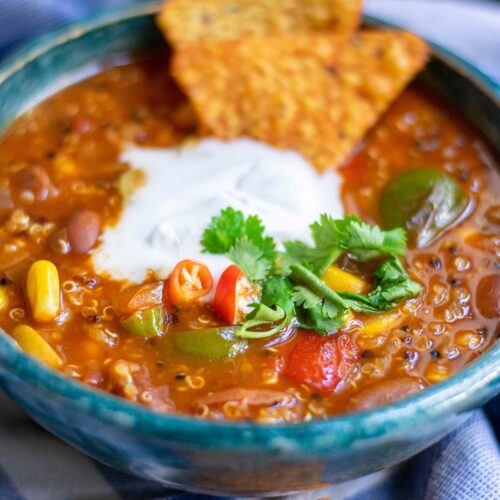 a bowl with quinoa chili, white cream on top, cilantro, and dorito chips on top of a blue towel.