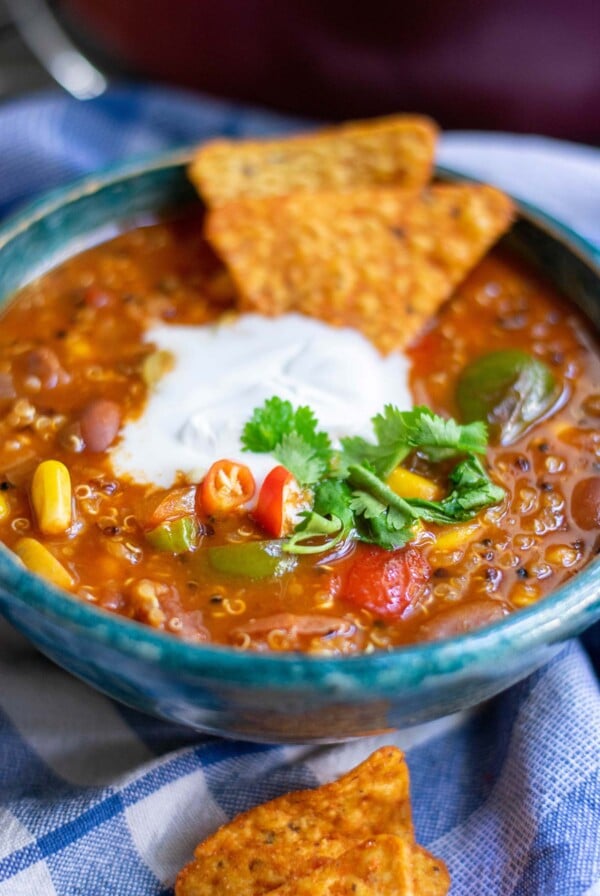 a bowl with quinoa chili, white cream on top, cilantro, and dorito chips on top of a blue towel.