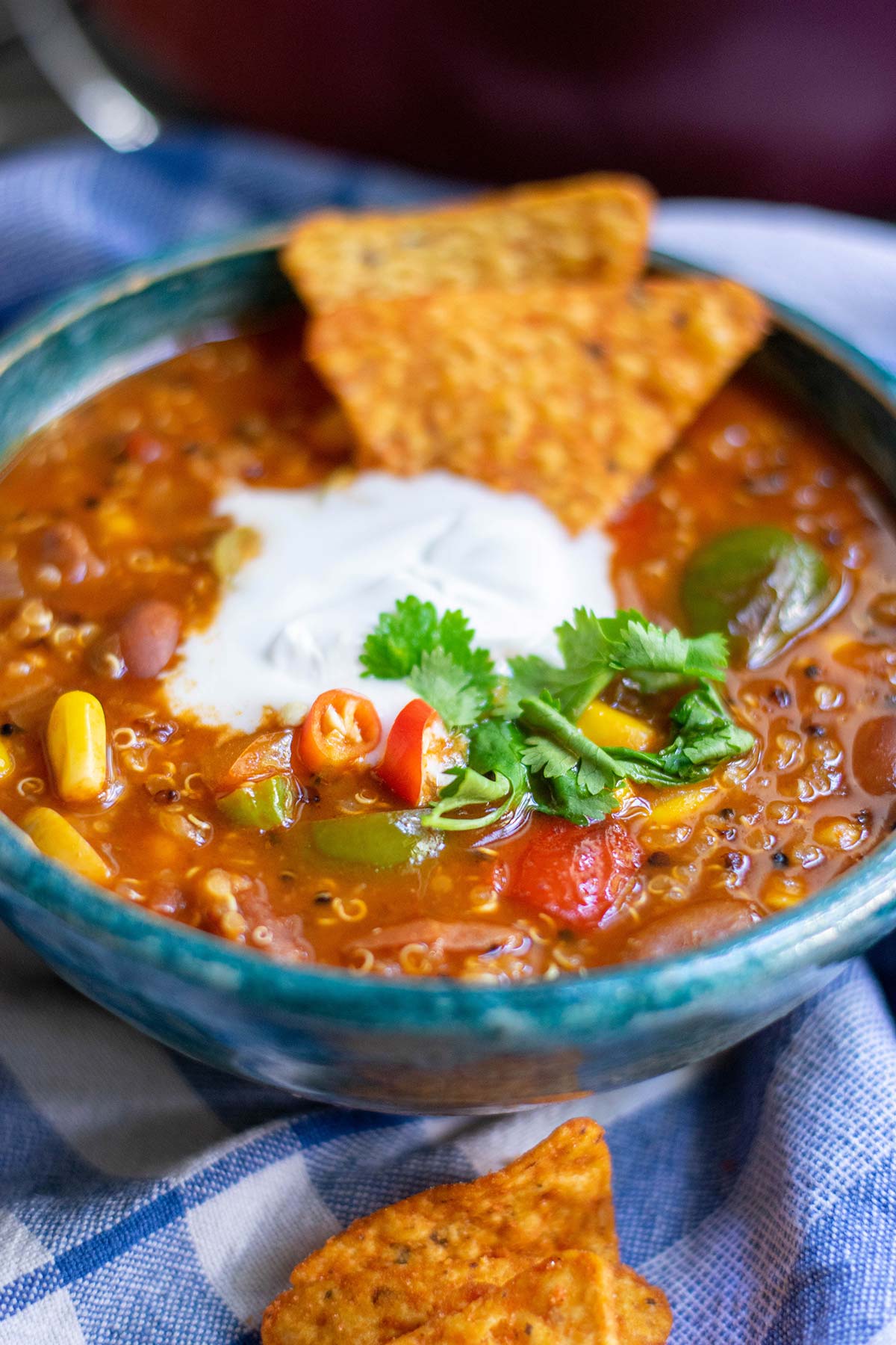 a bowl with quinoa chili, white cream on top, cilantro, and dorito chips on top of a blue towel. 