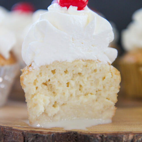 The inside of a tres leches cupcake with whipped cream and a cherry on top. Placed on a wooden surface.