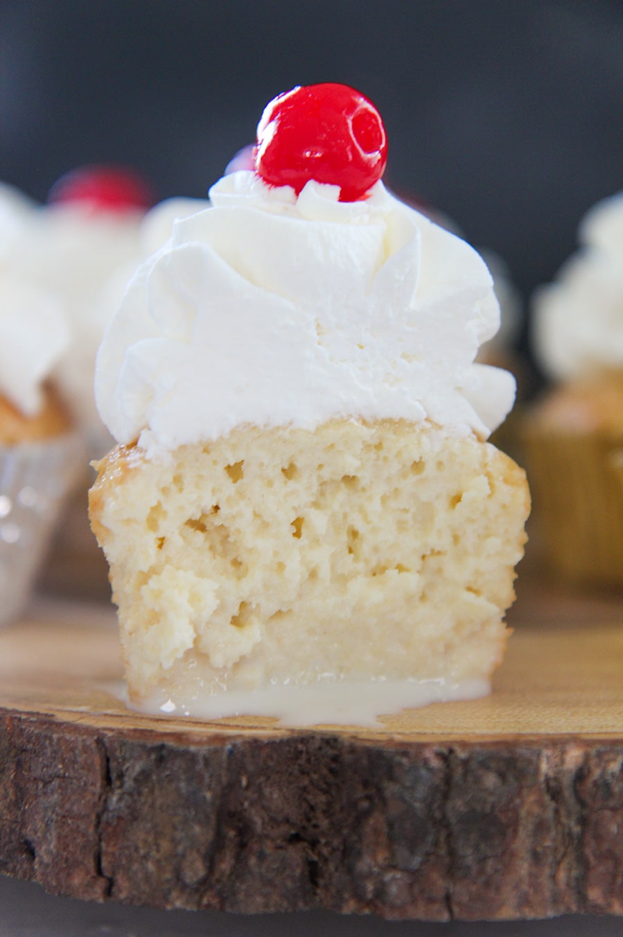 The inside of a tres leches cupcake with whipped cream and a cherry on top. Placed on a wooden surface. 