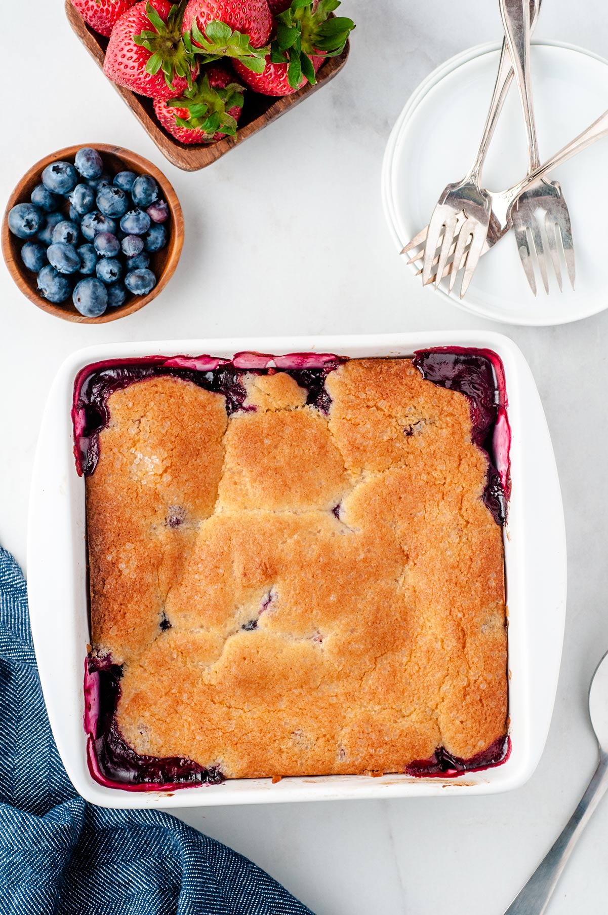berry cobbler in a white baking dish with strawberries and blueberries on the side. Forks and a small plate on the side. 